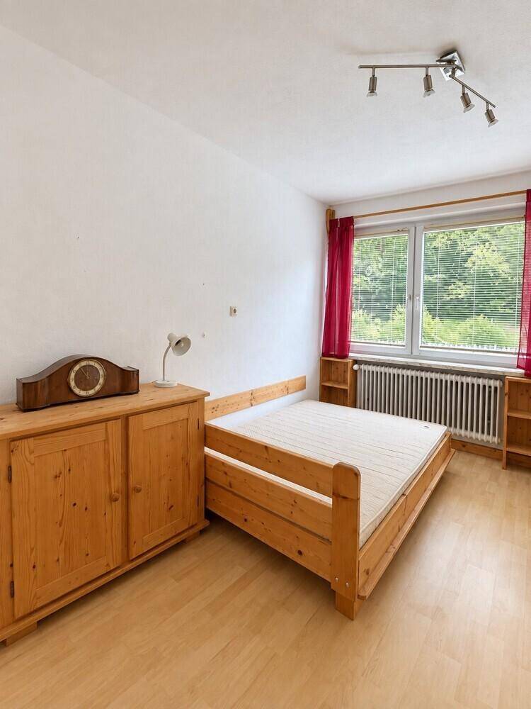 Helles Schlafzimmer mit Holzbett, Kommode und Fenster mit Blick ins Grüne.
