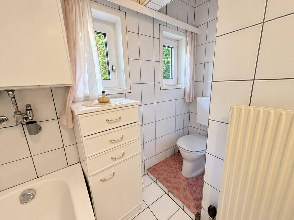 Badezimmer mit weißen Fliesen, Badewanne, Toilette und zwei kleinen Fenstern für Tageslicht.
