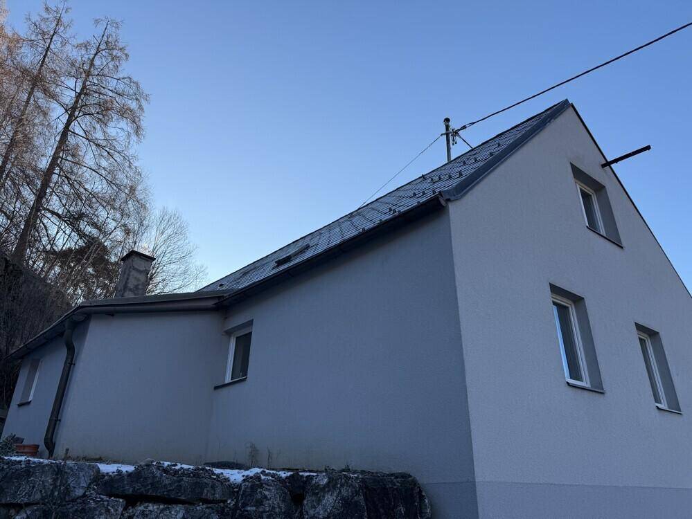 Rückansicht des Hauses mit Giebeldach und Schornstein vor einem bewaldeten Hang.
