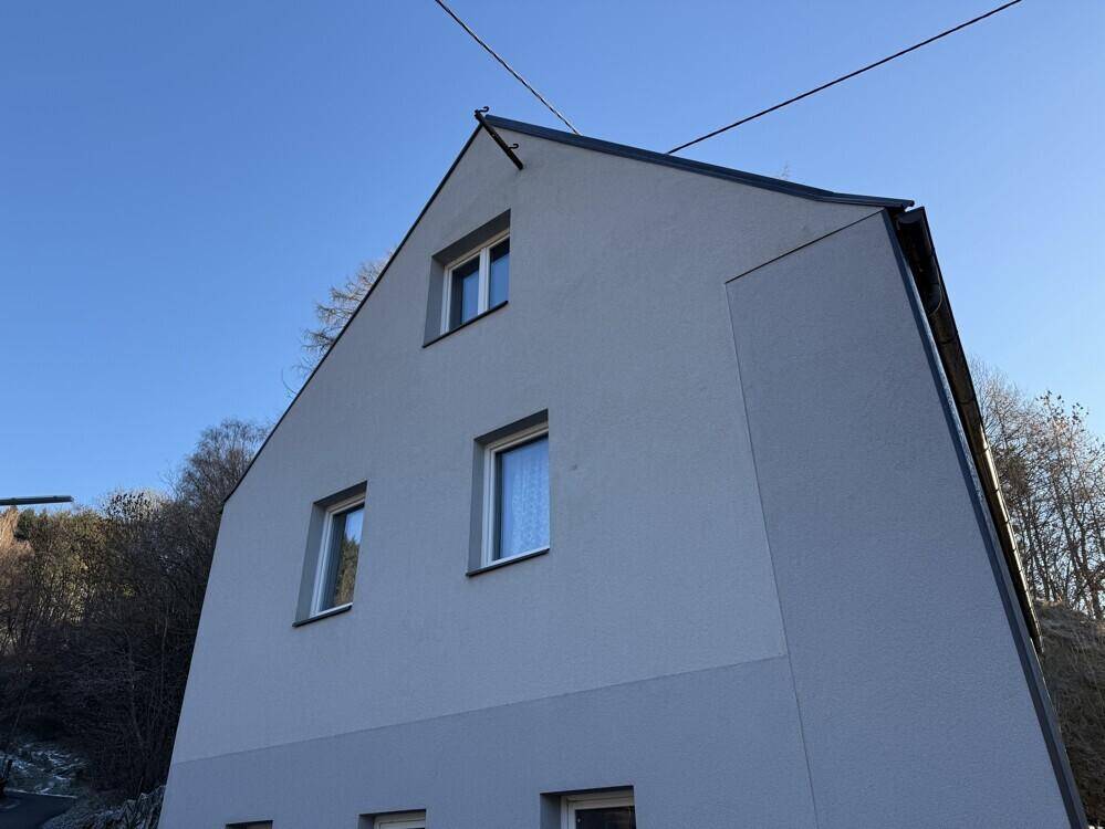 Seitenansicht des Hauses mit Giebeldach und mehreren Fenstern unter blauem Himmel.