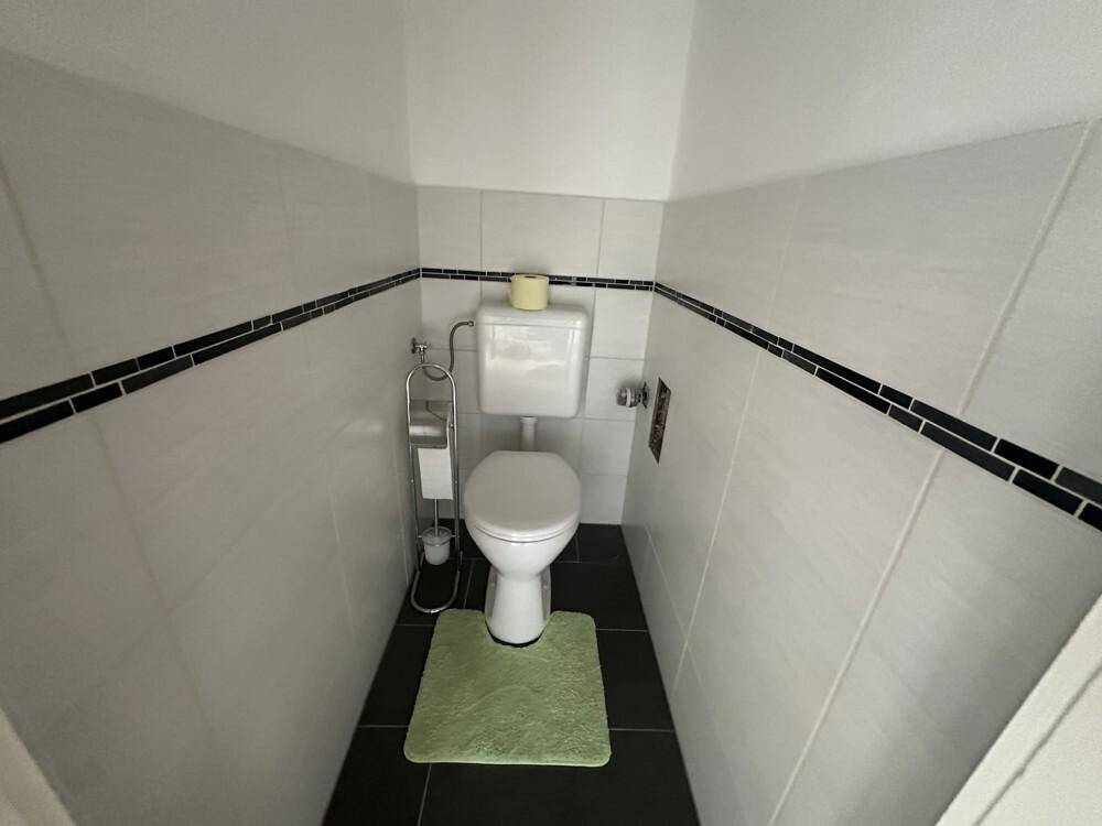 Separates WC mit weißen Fliesen, dunklem Boden und einer grünen Badematte.