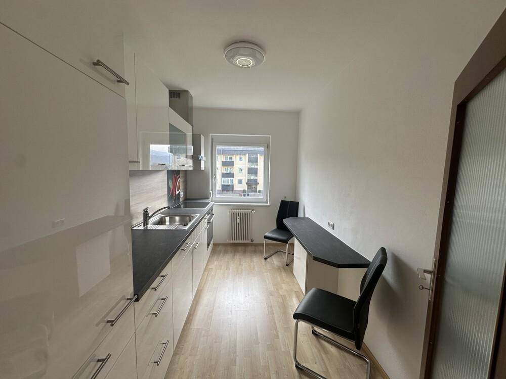 Moderne Küche mit weißen Hochglanzschränken, dunkler Arbeitsplatte und Essbereich am Fenster.