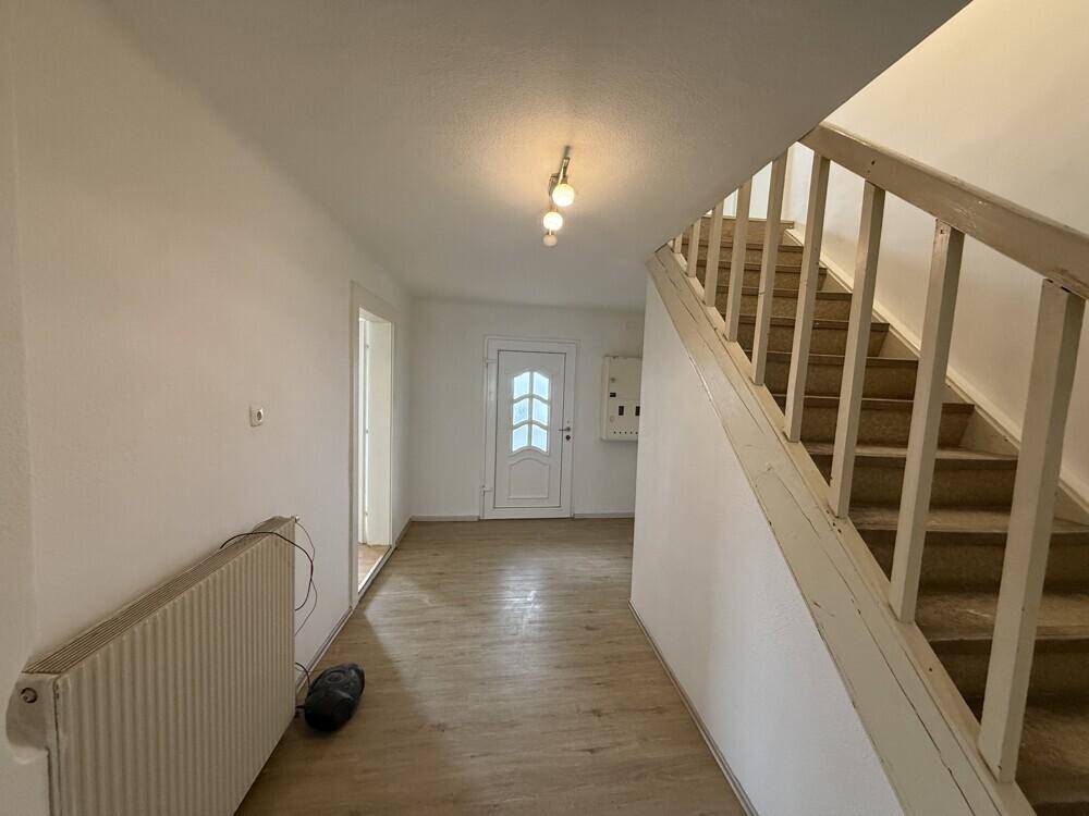 Heller Flur mit Holztreppe, Laminatboden und Zugang zur Eingangstür, ausgestattet mit Heizkörper.