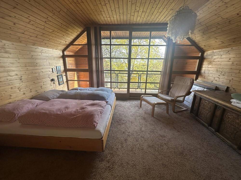 Gemütliches Schlafzimmer mit Holztäfelung, Teppichboden und großen Fenstern mit Blick ins Grüne.