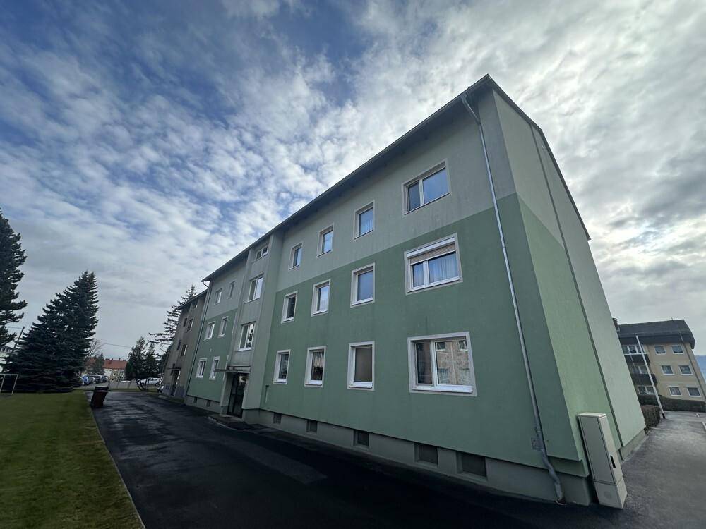Seitenansicht eines Mehrfamilienhauses mit grüner Fassade und Parkplatz.