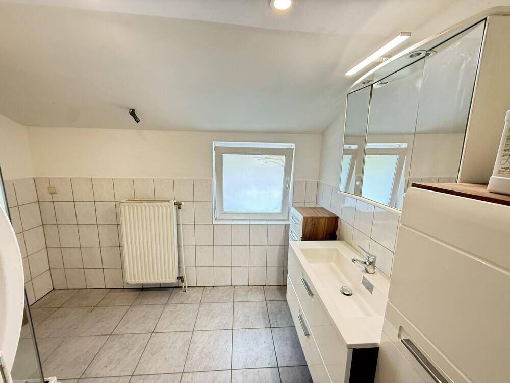 Geräumiges Badezimmer mit Waschtisch, Spiegelschrank, Fenster und weißen Fliesen an Wänden und Boden.
