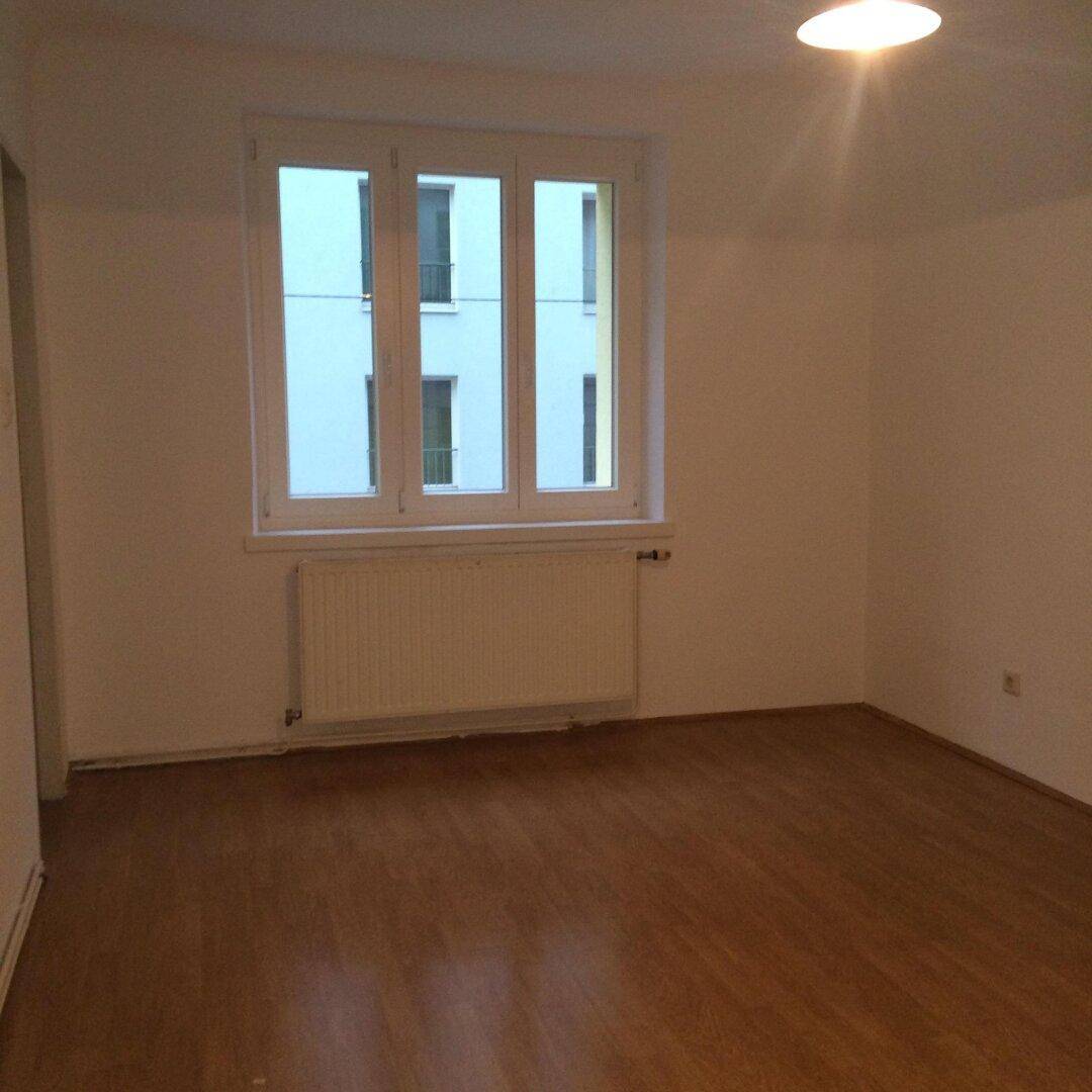 Unmöblierter Raum mit Holzfußboden und Fenster, das auf ein gegenüberliegendes Gebäude blickt.