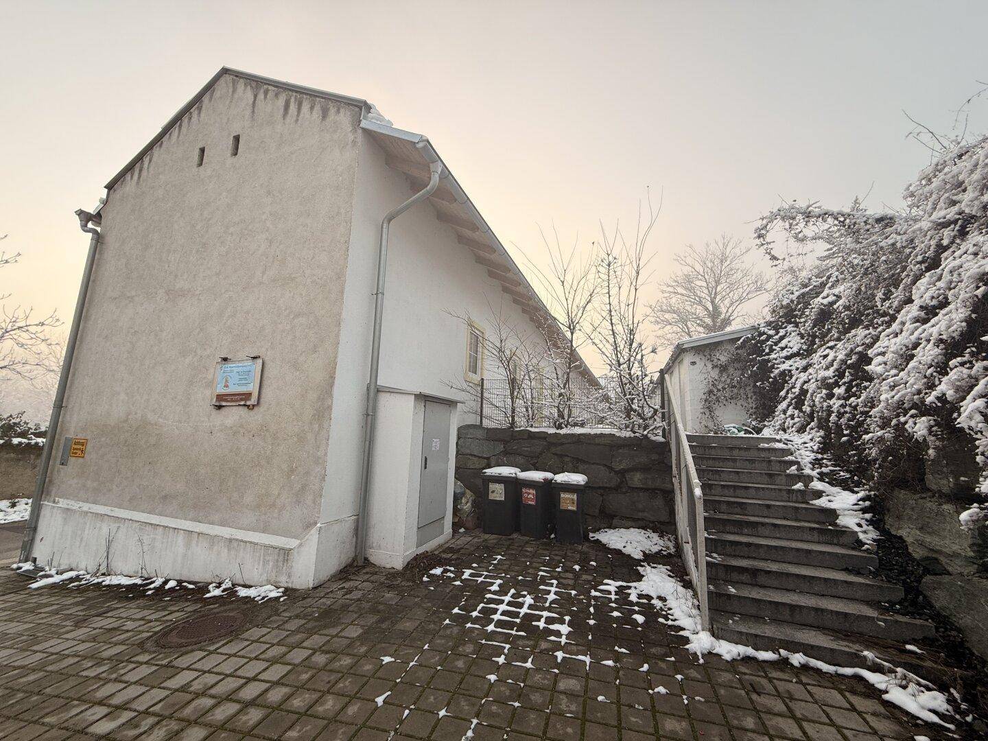 Gebäudeecke mit Außentreppe, Mülltonnen und verschneiter Umgebung.