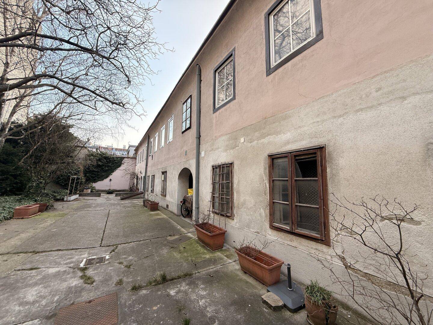 Langer, gepflasterter Innenhof eines Gebäudekomplexes mit Topfpflanzen und Bäumen.