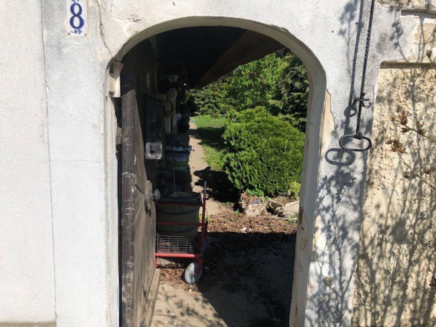 Blick durch einen gewölbten Torbogen auf einen sonnigen Gartenweg mit üppigem Grün.