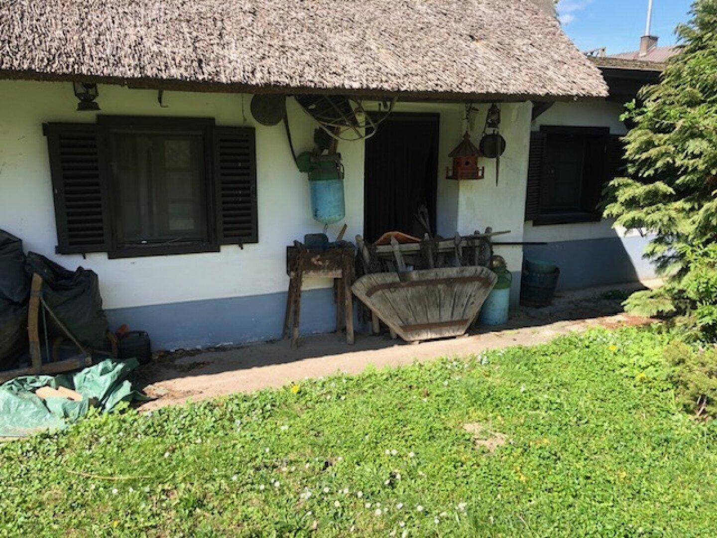 Seitenansicht des Hauses mit Fenstern, Fensterläden und Reetdach, davor verschiedene Gartenutensilien.