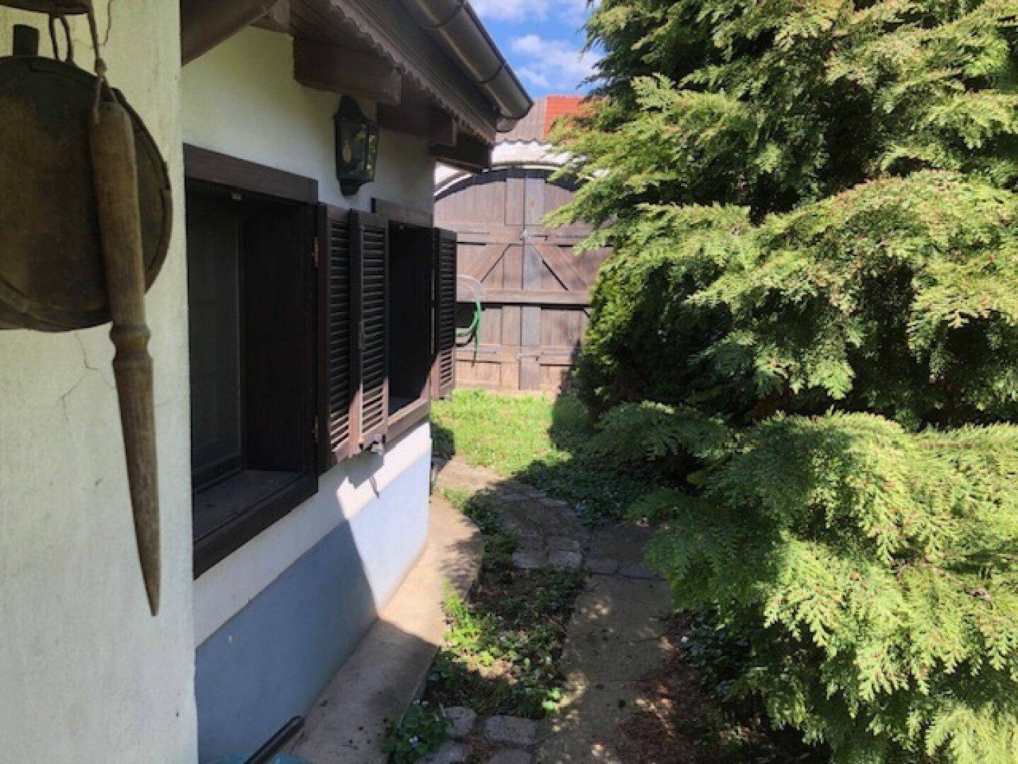 Schmaler Pfad entlang der Hauswand mit Fensterläden, der zu einem Holztor im grünen Garten führt.