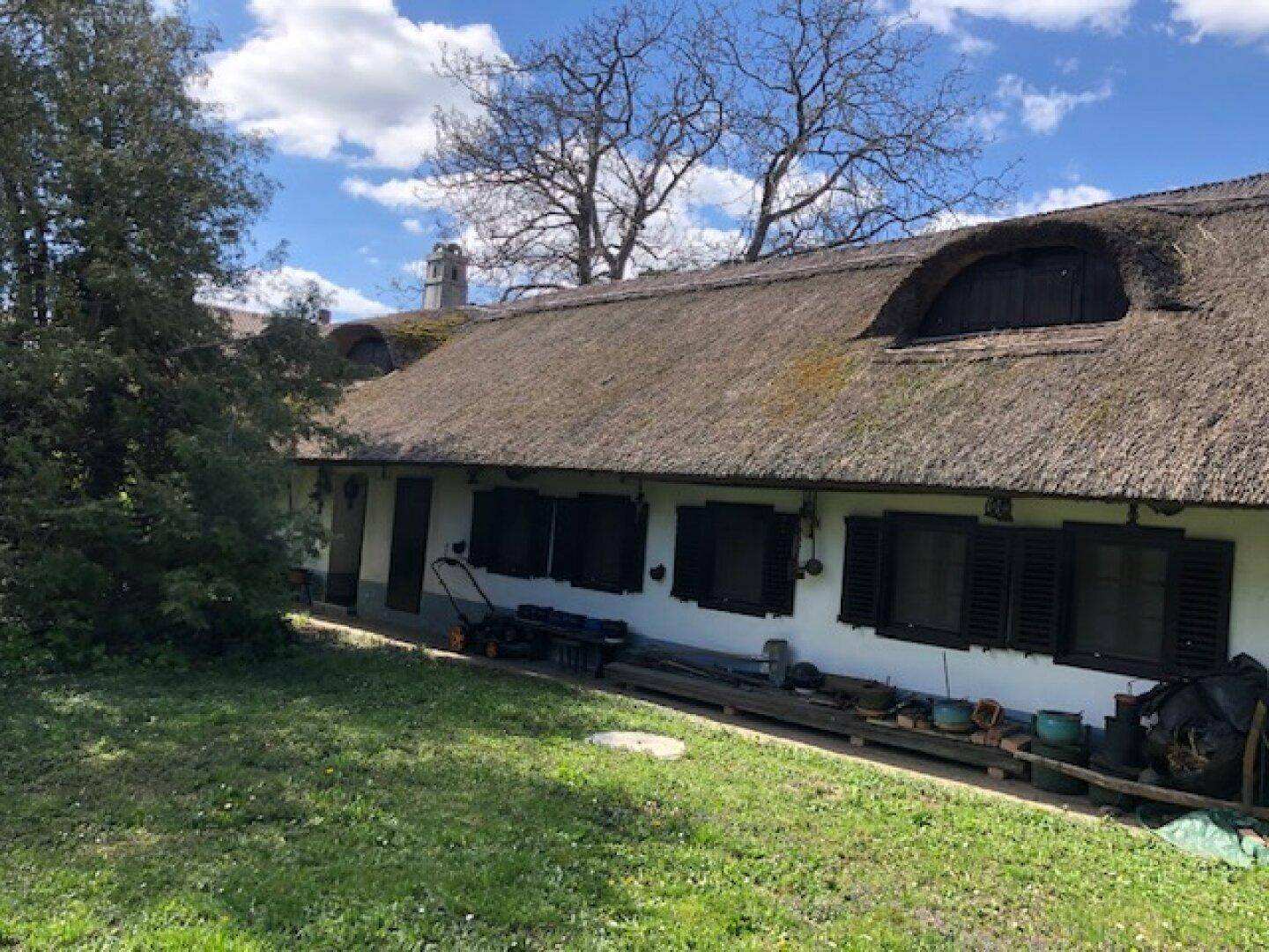 Ansicht des Hauses vom Garten aus, mit Reetdach, Fensterläden und einem Rasenmäher auf dem Weg.