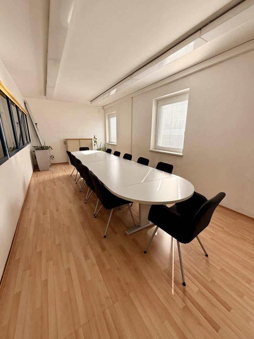 Großer Konferenzraum mit langem weißen Tisch, schwarzen Stühlen und hellem Laminatboden.