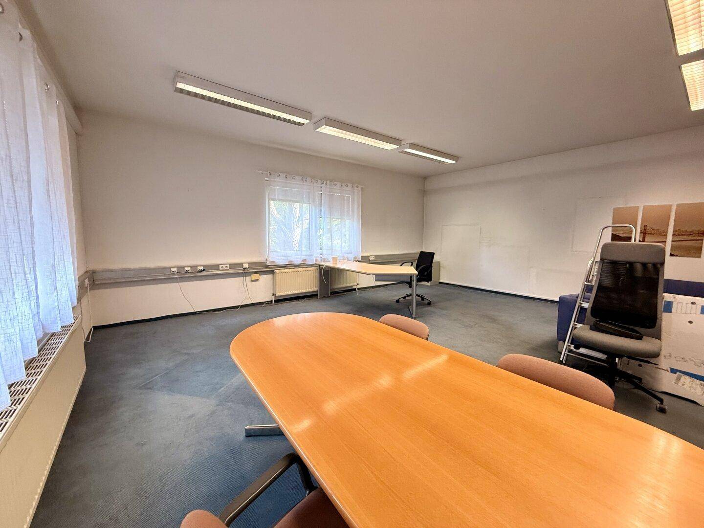 Büroraum mit großem Holztisch, blaugrauem Teppichboden und Fenstern mit hellen Vorhängen.
