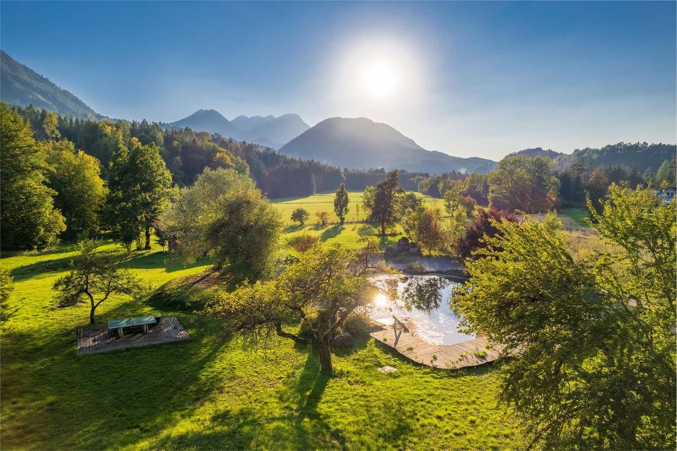 Idyllische Landschaft mit Teich, Holzdeck und Sitzbereich, umgeben von grünen Bäumen und Bergen.