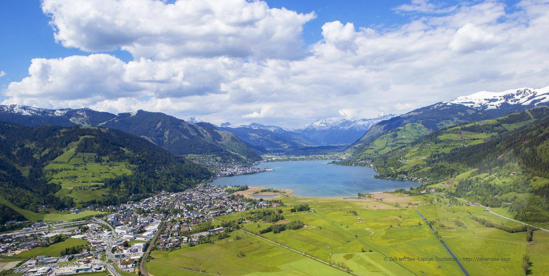 Panoramablick aus der Vogelperspektive auf einen malerischen See, umgeben von einer Stadt und grünen Bergen.