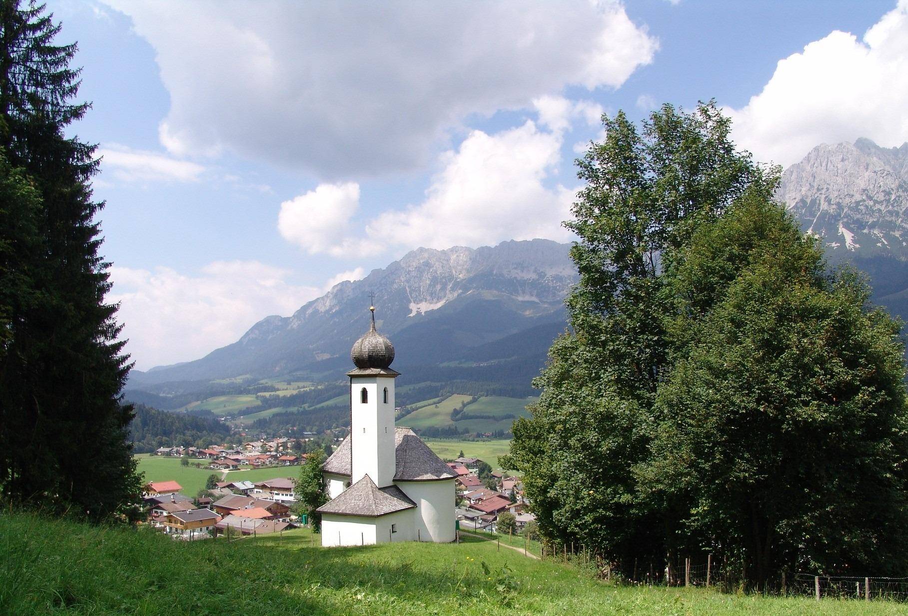 Idyllische Kirche mit Zwiebelturm in einem malerischen Dorf, umgeben von grünen Hügeln und Bergen.