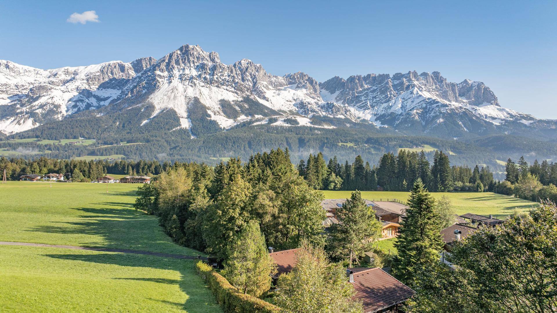 Weitläufige grüne Landschaft mit Häusern und Wäldern vor einer beeindruckenden Bergkulisse.