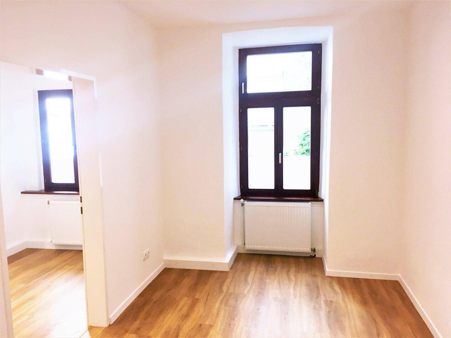 Leerer Raum mit Laminatboden, weißen Wänden und einem hohen Fenster, das den Raum erhellt.
