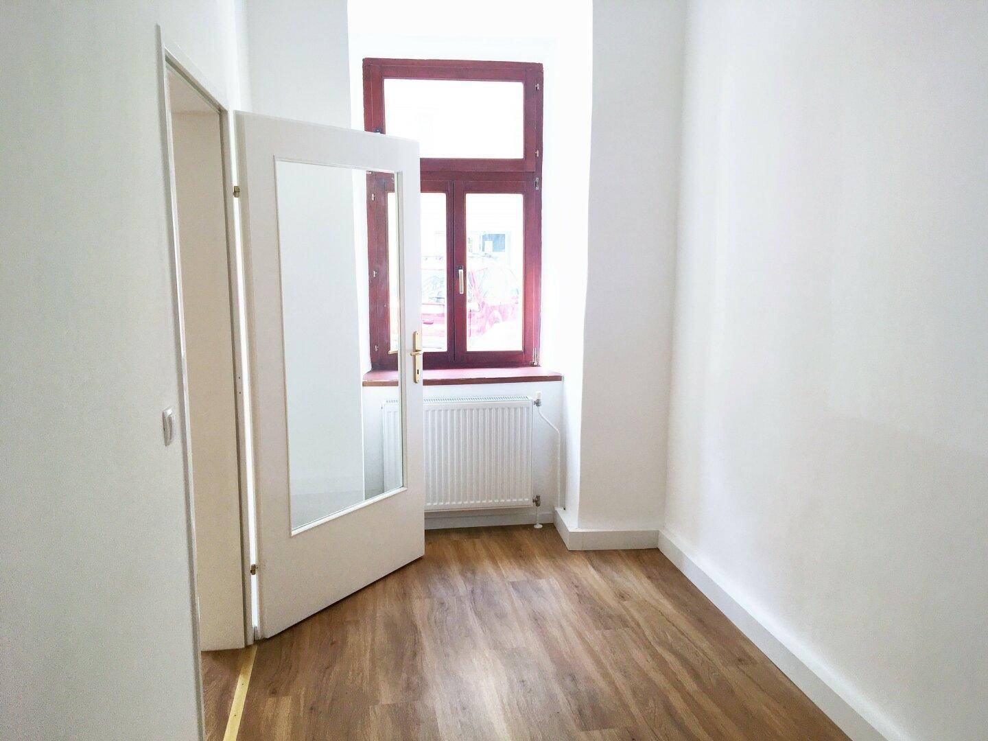 Heller, leerer Raum mit Laminatboden, weißen Wänden und einem Fenster, das den Raum mit Licht füllt.