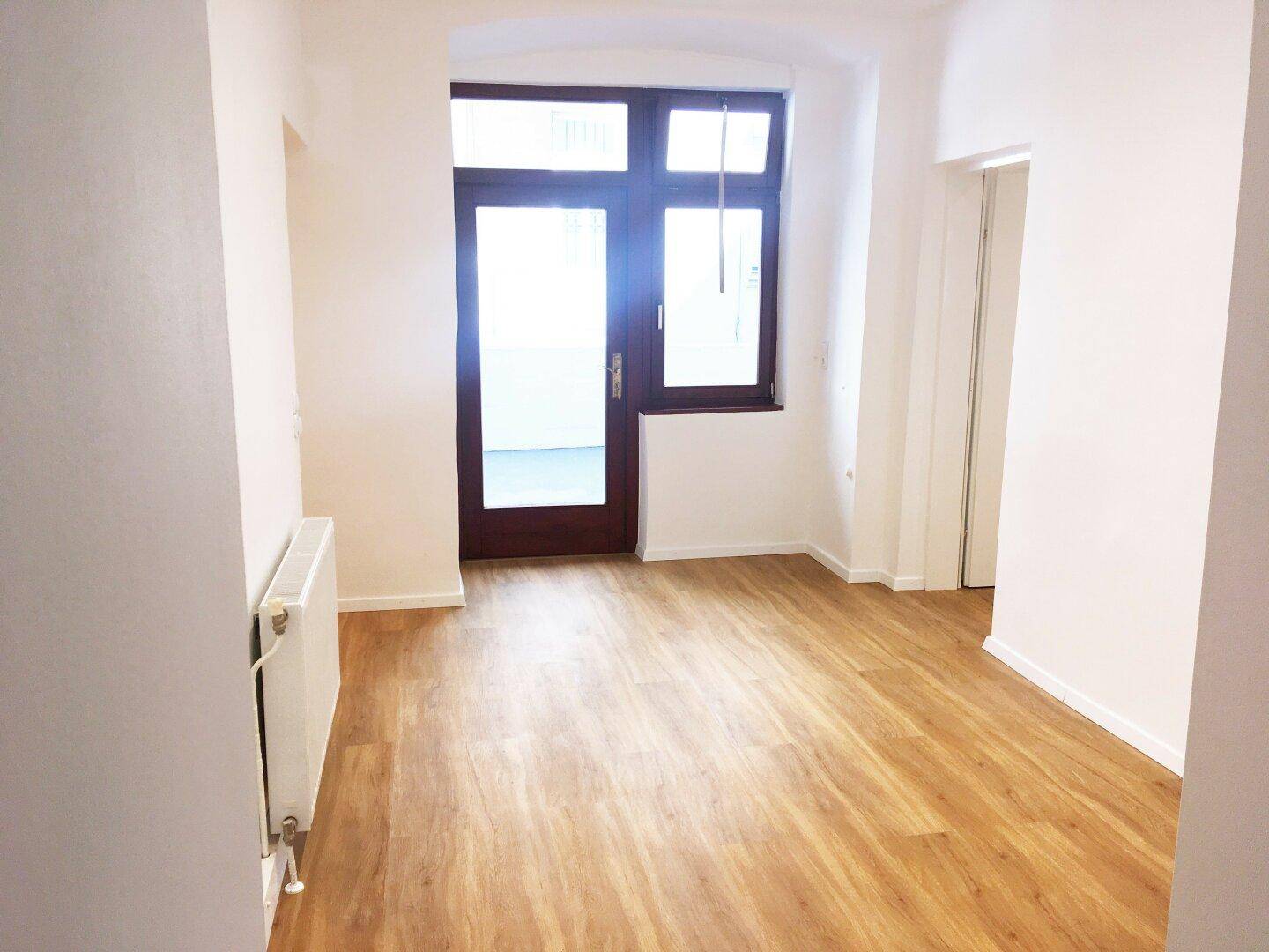 Heller Raum mit Laminatboden, weißen Wänden und einer Terrassentür, die Zugang zum Außenbereich bietet.