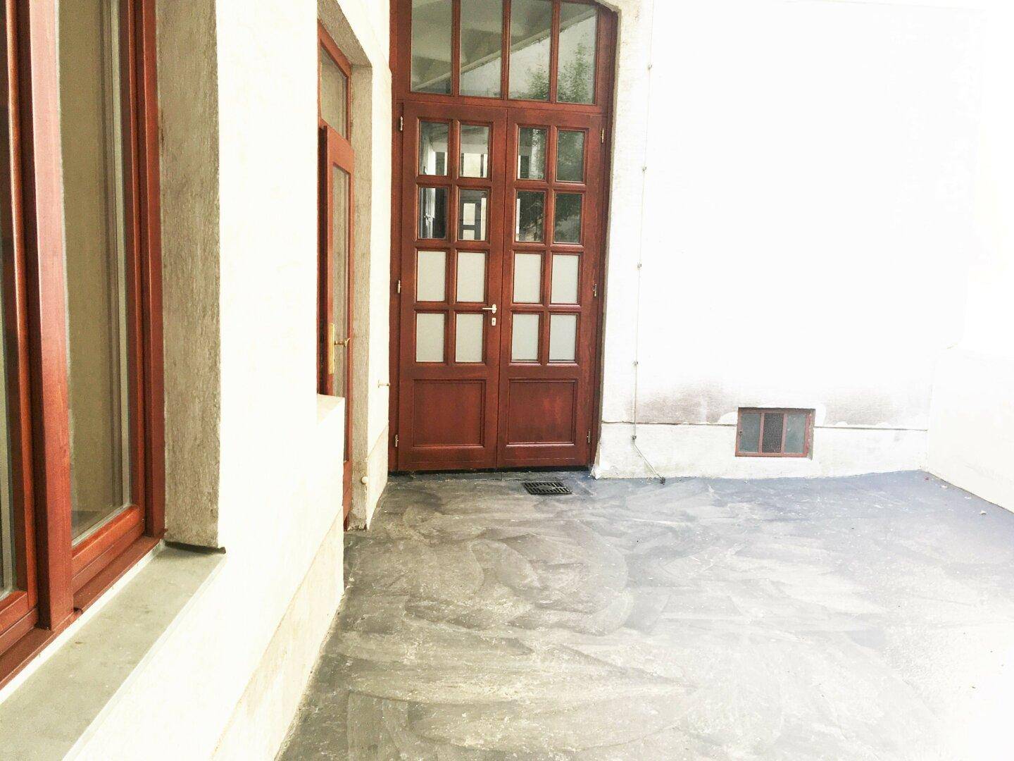 Große, gepflasterte Terrasse mit Holztüren, die zu den Innenräumen des Gebäudes führen.