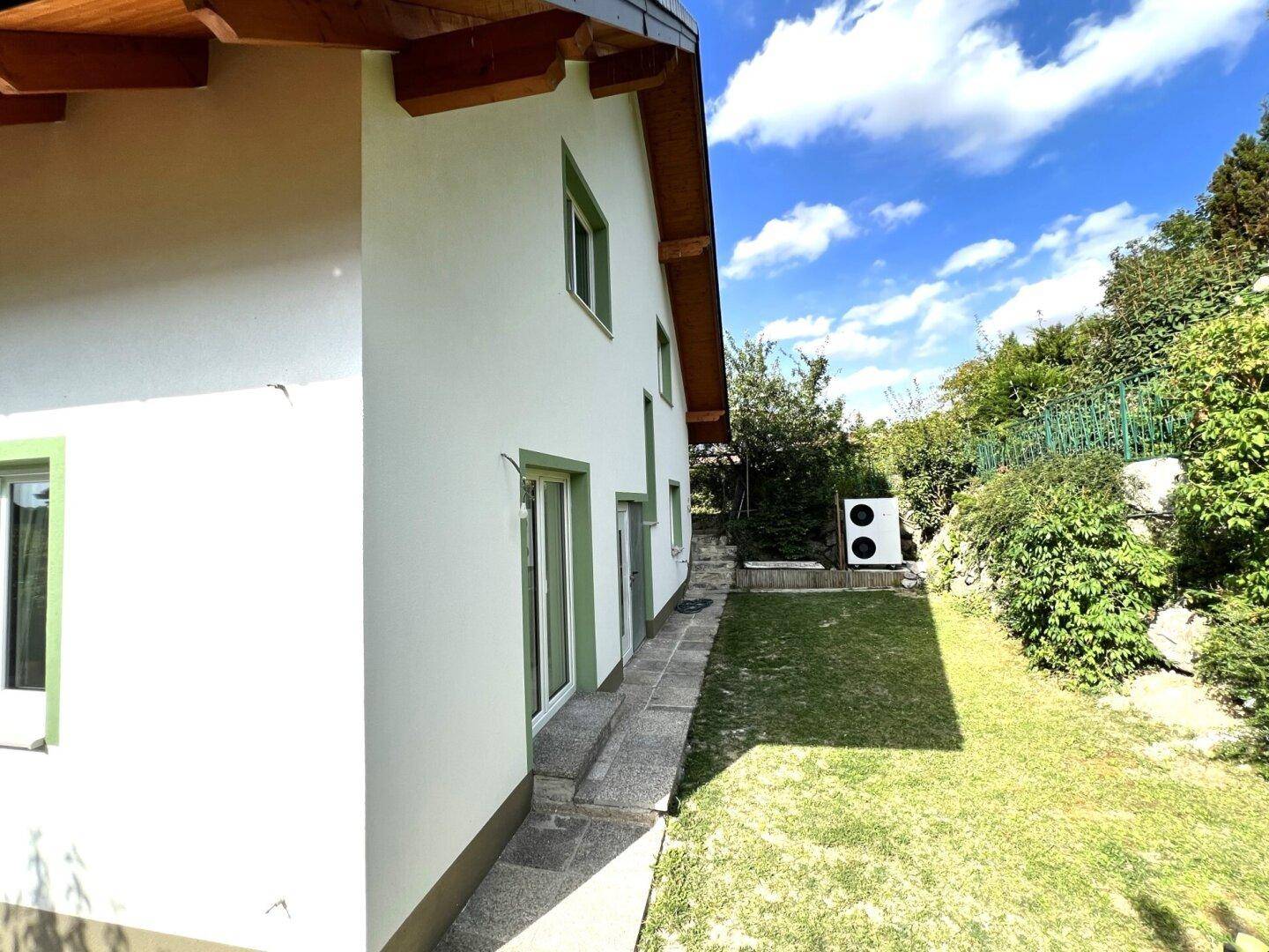 Gepflegter Rasenbereich neben dem Haus mit einer Wärmepumpe und üppiger Bepflanzung im Hintergrund.