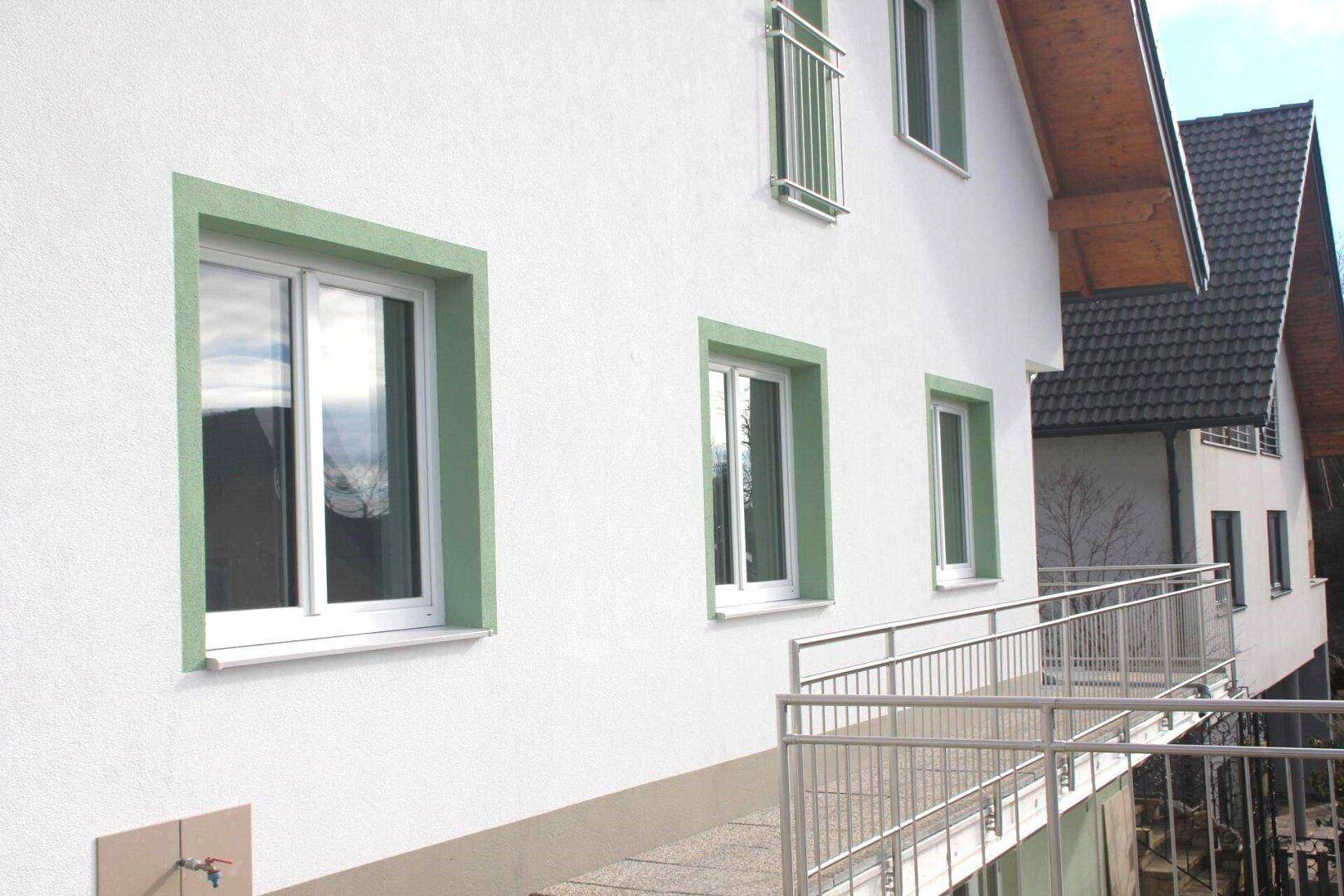 Detailansicht der weißen Fassade mit grünen Fensterrahmen und einem modernen Metallgeländer.