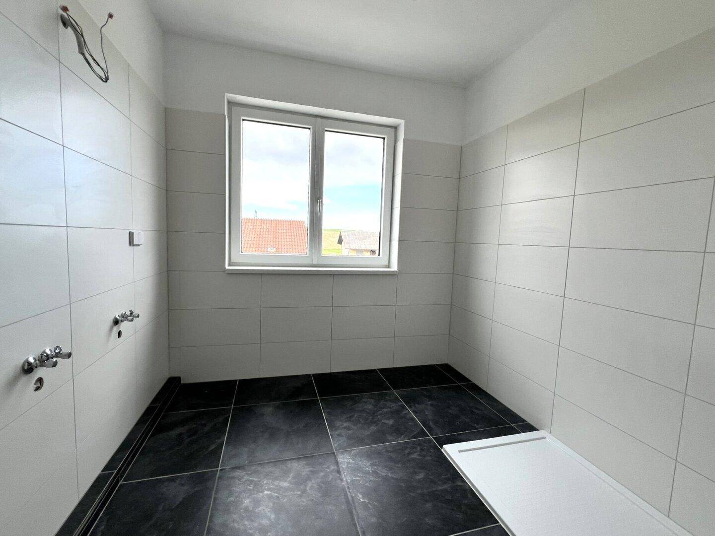 Unfertiges Badezimmer mit weißen Wandfliesen, dunklen Bodenfliesen und einem Fenster.