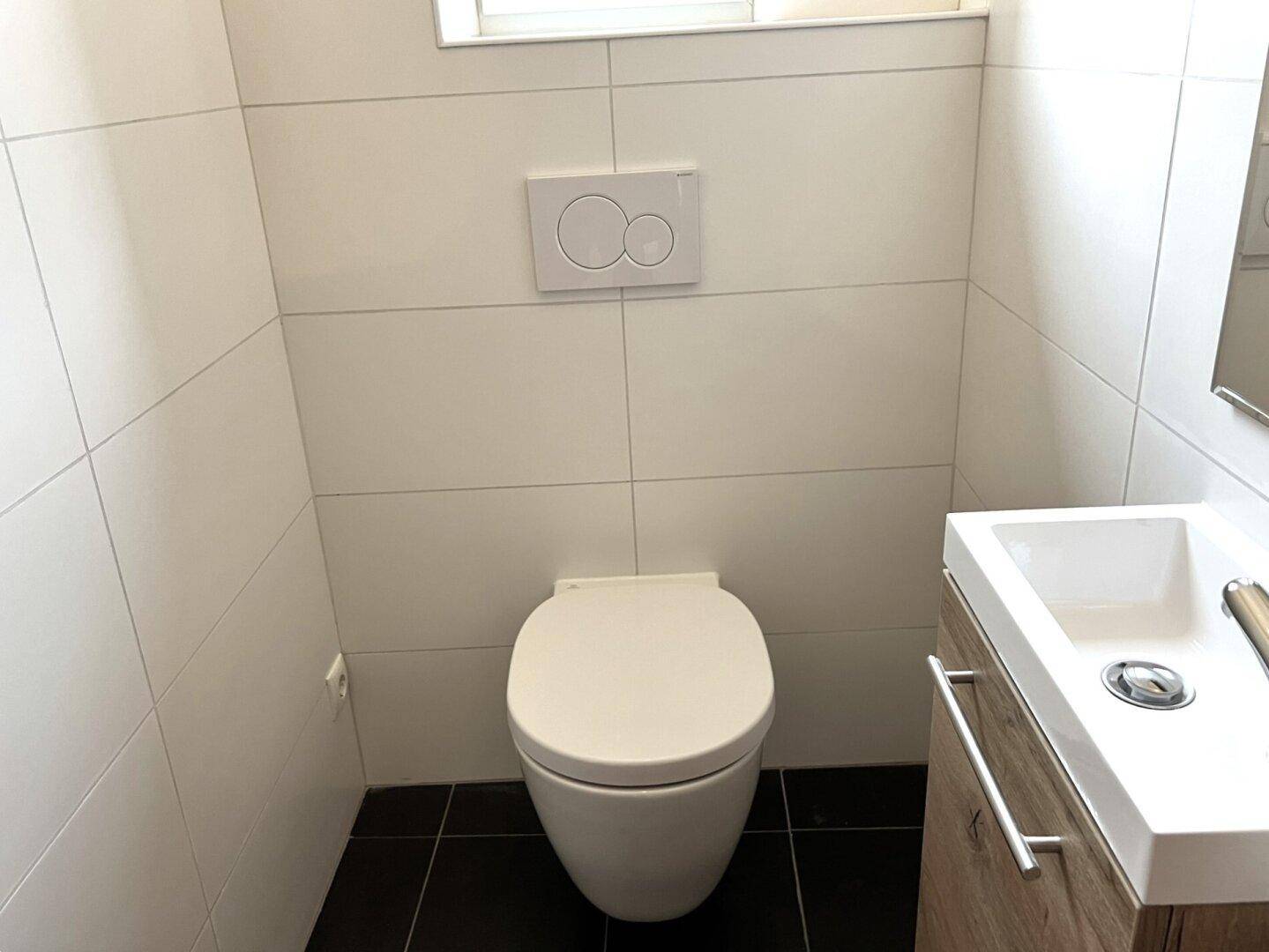 Modernes Gäste-WC mit weißen Fliesen, dunklem Boden, wandhängendem WC und kleinem Waschtisch.