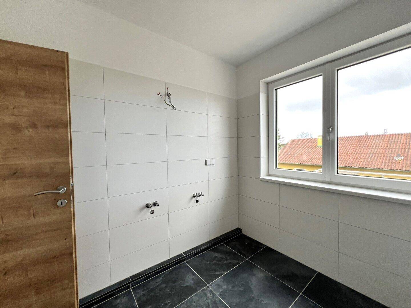 Unfertiges Badezimmer mit weißen Wandfliesen, dunklen Bodenfliesen und einem Fenster.