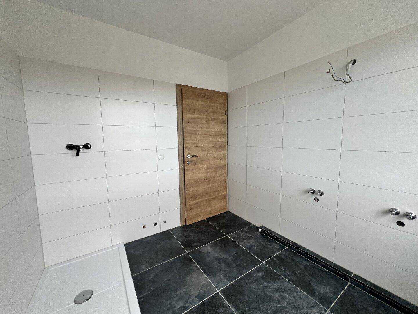 Unfertiges Badezimmer mit weißen Wandfliesen, dunklen Bodenfliesen und einer Holztür.