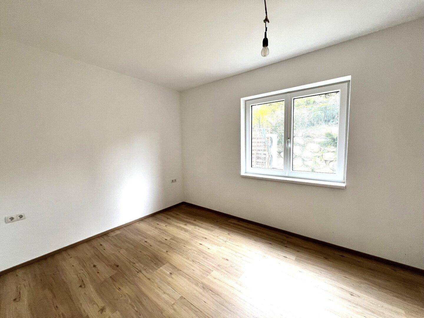 Helles, leeres Zimmer mit Laminatboden und einem Fenster mit Blick ins Grüne.