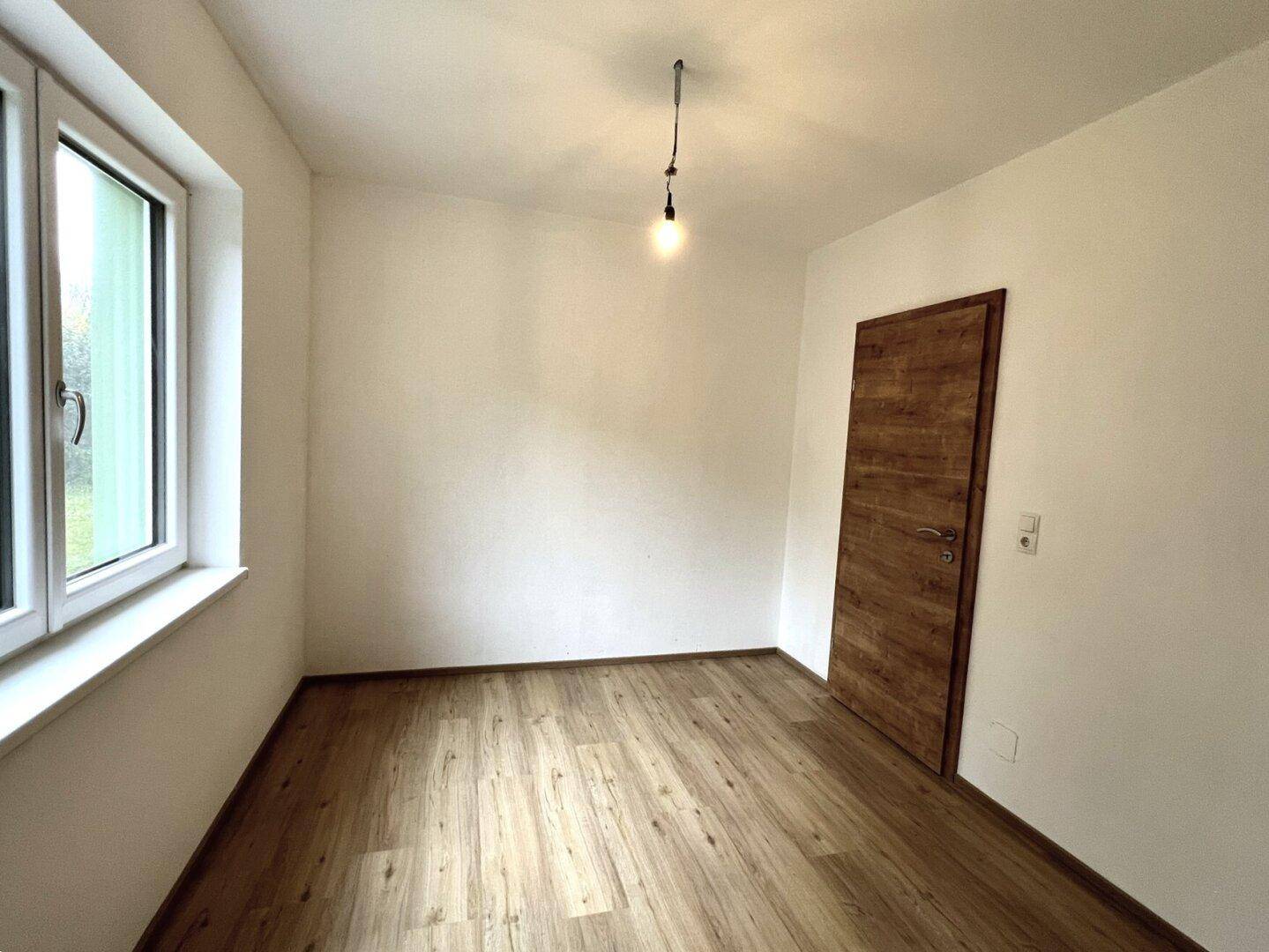 Helles, leeres Zimmer mit Laminatboden, weißen Wänden und einem Fenster.