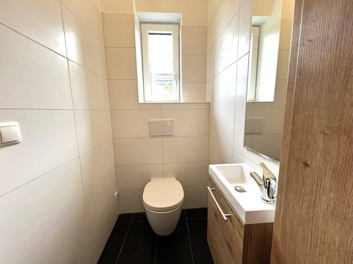 Helles Gäste-WC mit weißen Fliesen, dunklem Boden, wandhängendem WC und kleinem Waschtisch.