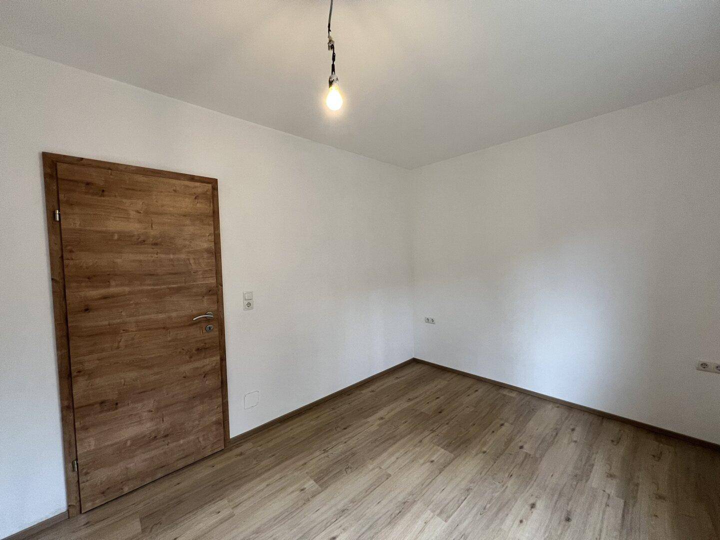 Leeres Zimmer mit Laminatboden, weißen Wänden und einer modernen Holztür.
