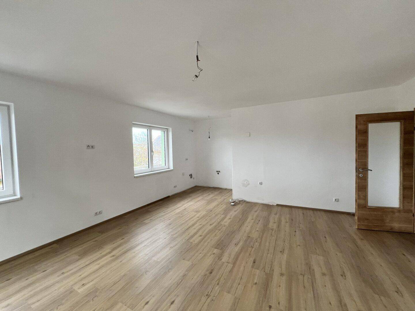 Großes, leeres Zimmer mit Laminatboden, mehreren Fenstern und einer Holztür.