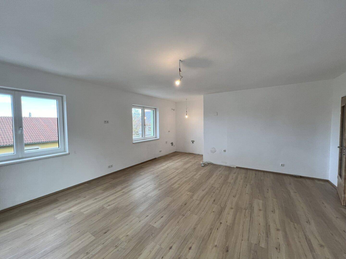 Großes, leeres Zimmer mit Laminatboden, mehreren Fenstern und einer Holztür.