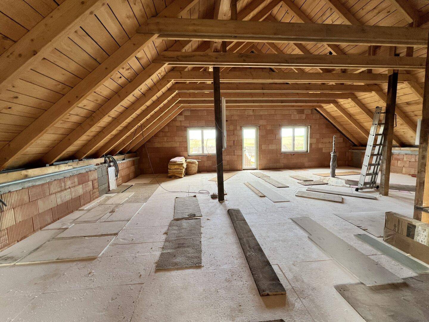Unausgebauter Dachboden mit sichtbaren Holzbalken, Ziegelwänden und Fenstern, bereit für den Ausbau.