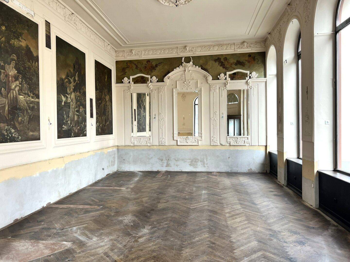 Historischer Raum mit Fischgrätparkett, hohen Decken, Wandgemälden und dekorativen Spiegelelementen, bereit zur Renovierung.