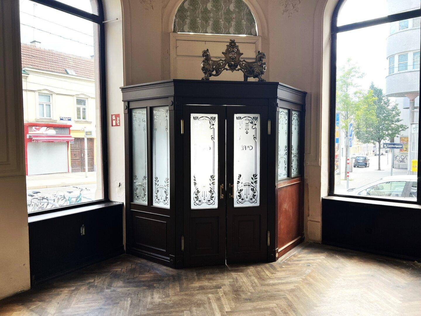 Historischer Eingangsbereich mit verzierten Glastüren und Blick auf die Straße, umgeben von hohen Fenstern.