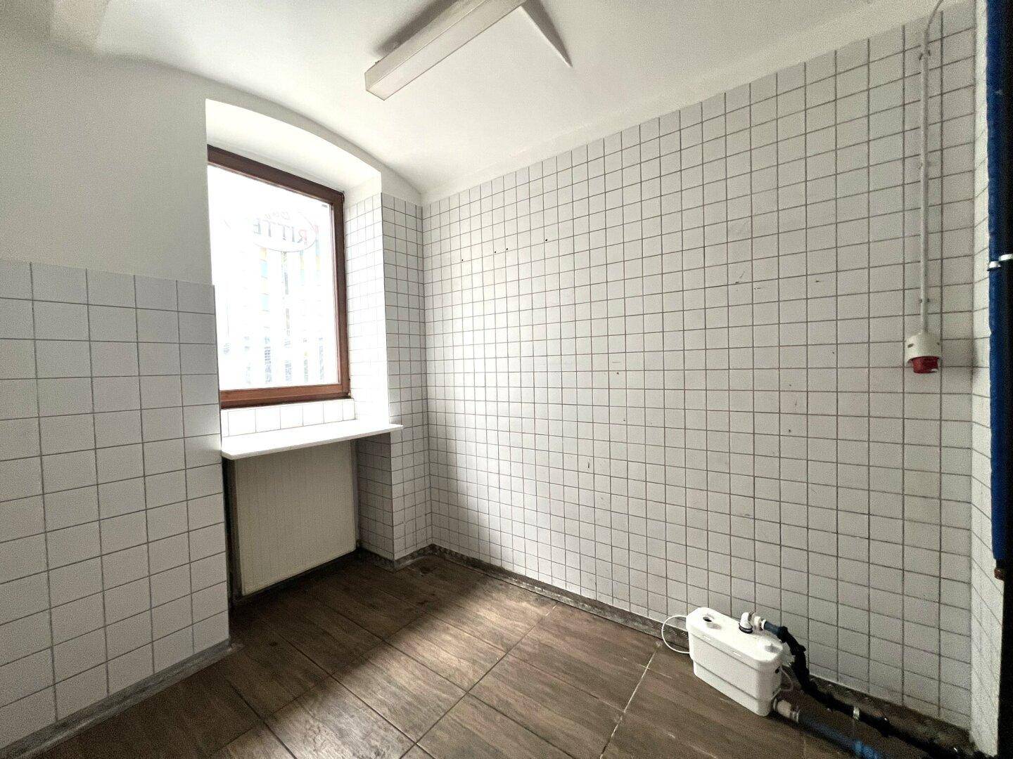 Kleiner Raum mit Fliesenwänden, einem Fenster und sichtbaren Installationen, der renoviert werden muss.