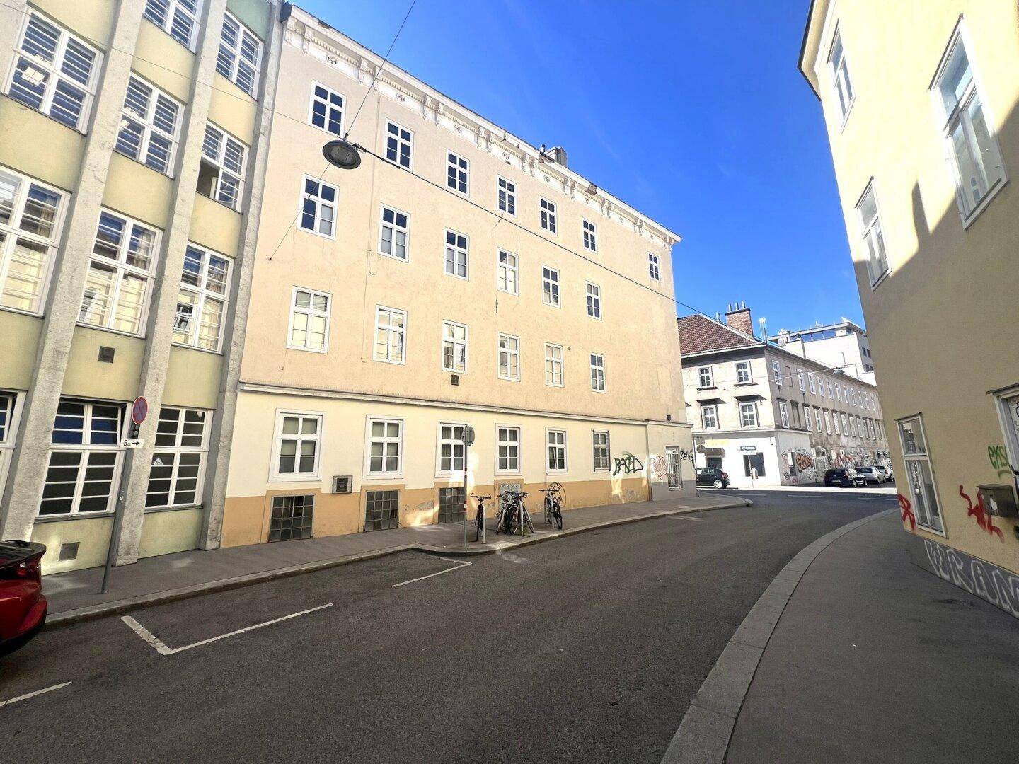 Seitenansicht eines Mehrfamilienhauses in einer sonnigen Stadtstraße mit Fahrrädern und geparkten Autos.