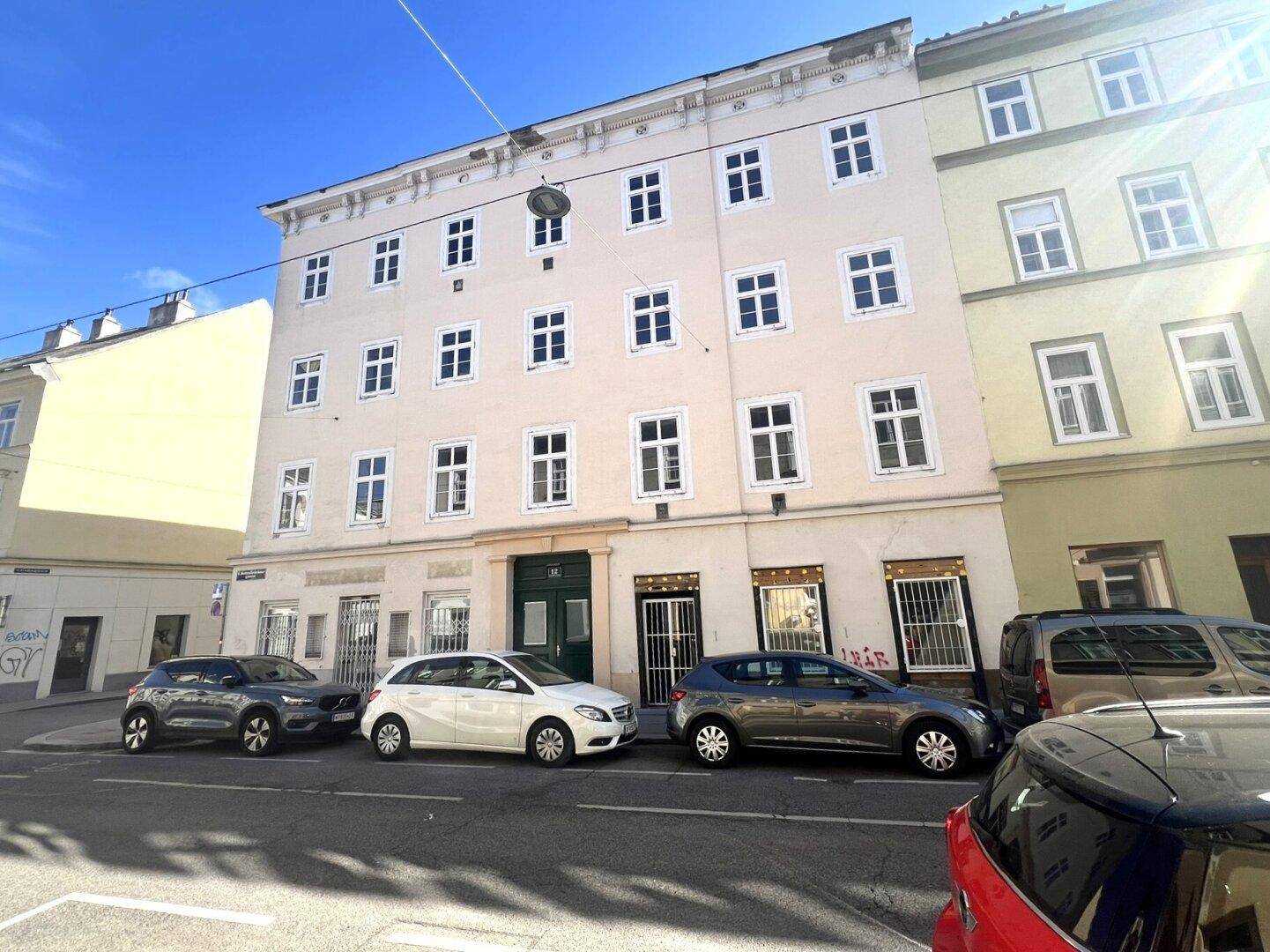 Vorderansicht eines klassischen Mehrfamilienhauses mit vielen Fenstern und geparkten Fahrzeugen.