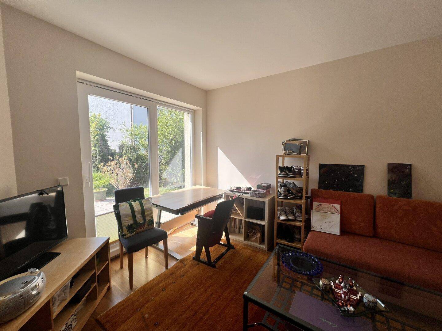 Heller Wohnbereich mit Sofa, Schreibtisch und großem Fenster zum Außenbereich.