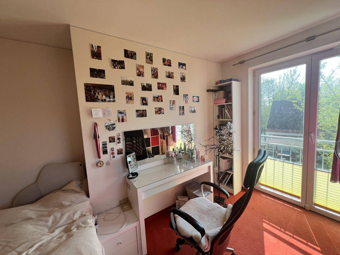 Schlafzimmer mit rotem Teppichboden, Schreibtisch und Zugang zum Balkon.