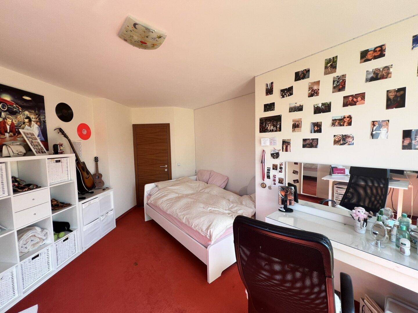 Schlafzimmer mit rotem Teppichboden, Bett und Schreibtisch, hell und freundlich.