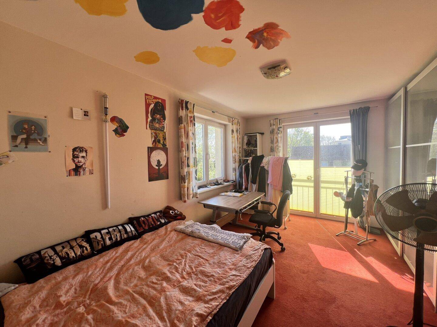Helles Schlafzimmer mit rotem Teppichboden, Bett und Schreibtisch, mit Balkonzugang.