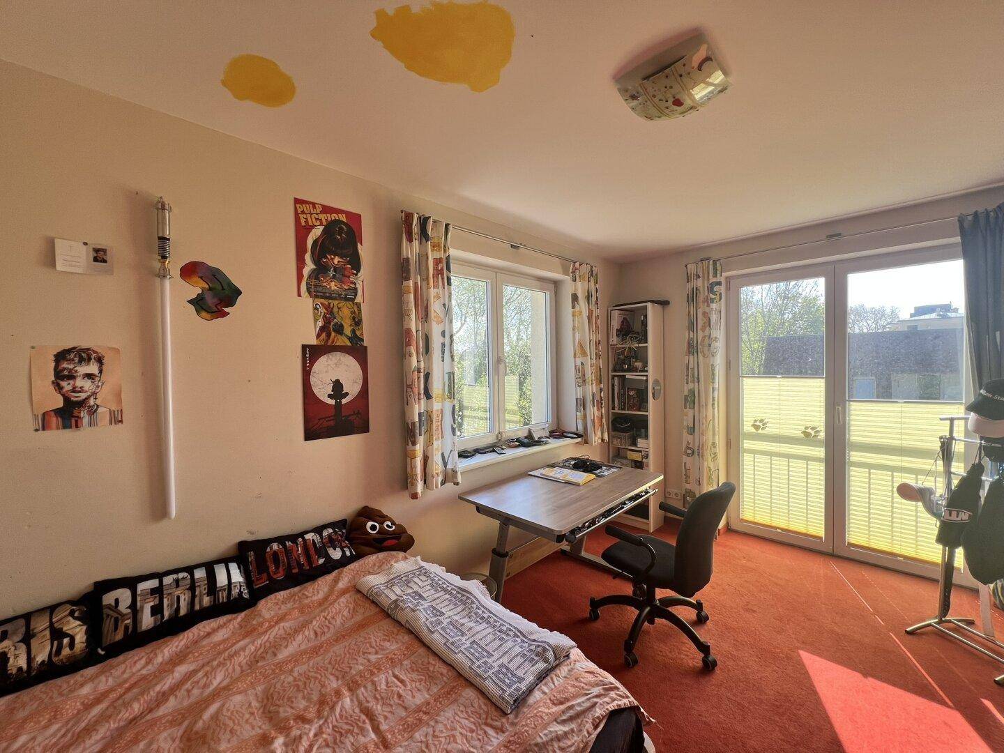 Schlafzimmer mit rotem Teppichboden, Schreibtisch und Fenster mit Blick ins Grüne.